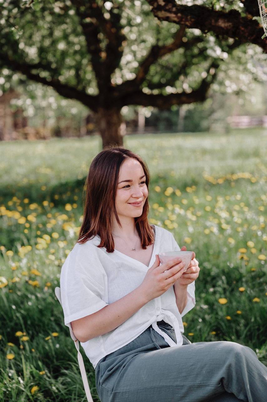 Junge Frau mit braunen, schulterlangen Haaren sitzt entspannt auf einem Stuhl in einer blühenden Wiese mit vielen gelben Blumen. Sie trägt eine weiße Bluse und graugrüne Hose und hält eine Tasse in beiden Händen. Im Hintergrund steht ein großer blühender Baum, der Schatten spendet. Die Szene wirkt ruhig und naturverbunden.
