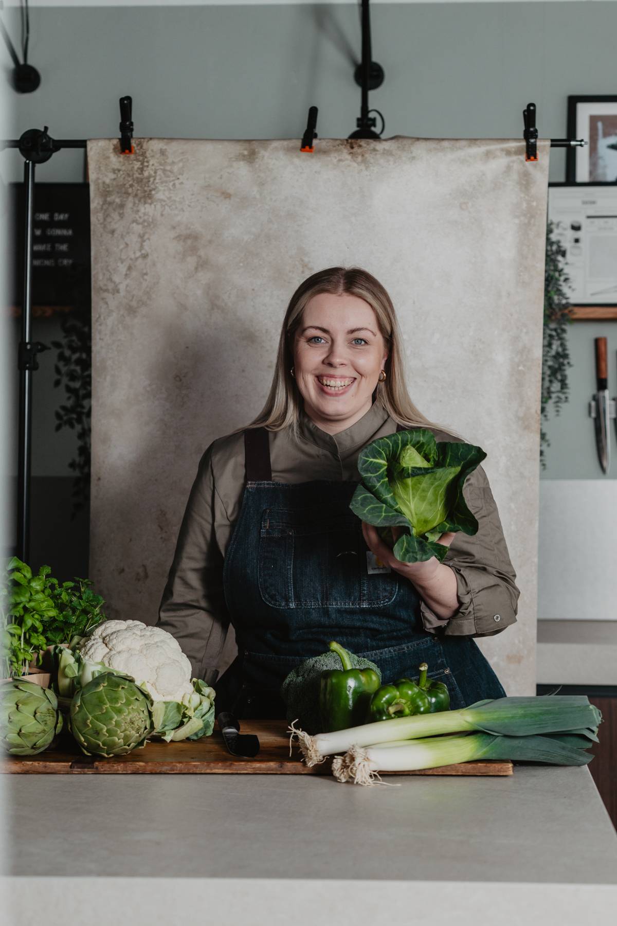 Junge blonde Frau steht in einer Küche und präsentiert frisches grünes Gemüse